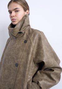 Γυναίκα που φοράει υπερμεγέθη δερμάτινο μπουφάν ταμπά με μαύρα κουμπιά και ψηλό γιακά, ποζάρει μπροστά σε απλό φωτεινό φόντο.