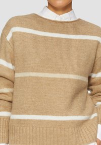 Beżowy sweter dziany z białymi poziomymi paskami. Prążkowana tekstura, okrągły dekolt i opadające ramiona. Nosi się go na białej koszuli z kołnierzykiem.