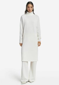 Weißes gestricktes Kleid mit hohem Rippenkragen, langen Ärmeln und geripptem Saum. Kombiniert mit weißen, weit geschnittenen Strickhosen.