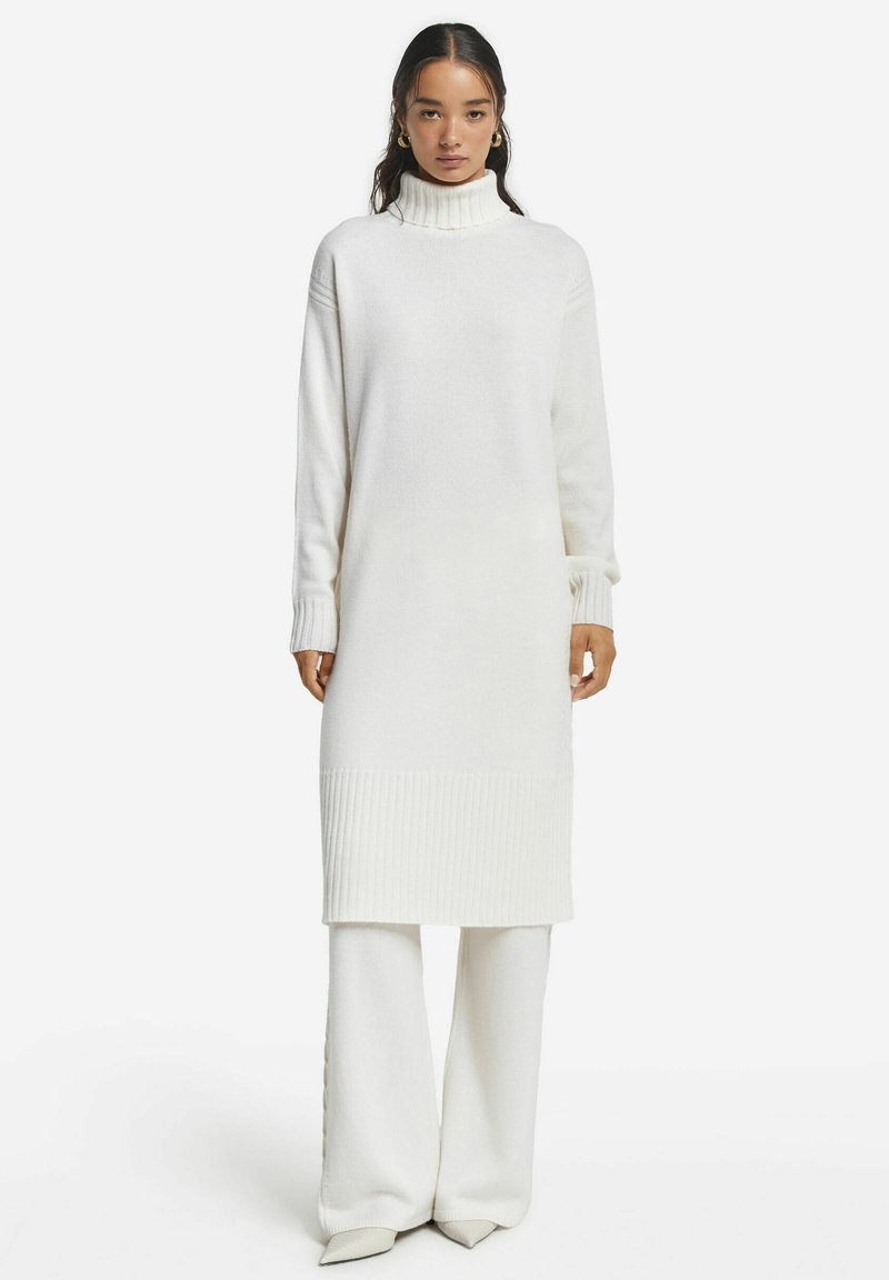 Weißes gestricktes Kleid mit hohem Rippenkragen, langen Ärmeln und geripptem Saum. Kombiniert mit weißen, weit geschnittenen Strickhosen.