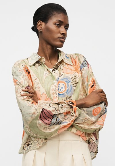 Femme aux cheveux foncés en chignon, portant une chemise à manches longues à motif floral et un pantalon plissé crème, bras croisés, regardant sur le côté.