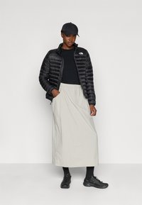 Black padded jacket, black top, light gray skirt with pockets, black socks, and shoes. Cap worn, standing against a neutral background.