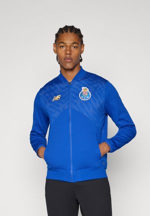 Chaqueta deportiva azul con cremallera y detalles de malla. Presenta un logo en el lado izquierdo del pecho y un dobladillo acanalado. Diseñada en una forma ajustada con mangas largas.