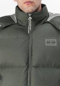 Giacca puffer verde con colletto alto, chiusura a zip e accenti grigi. Presenta un motivo a griglia e il logo sul petto. Materiale leggero.