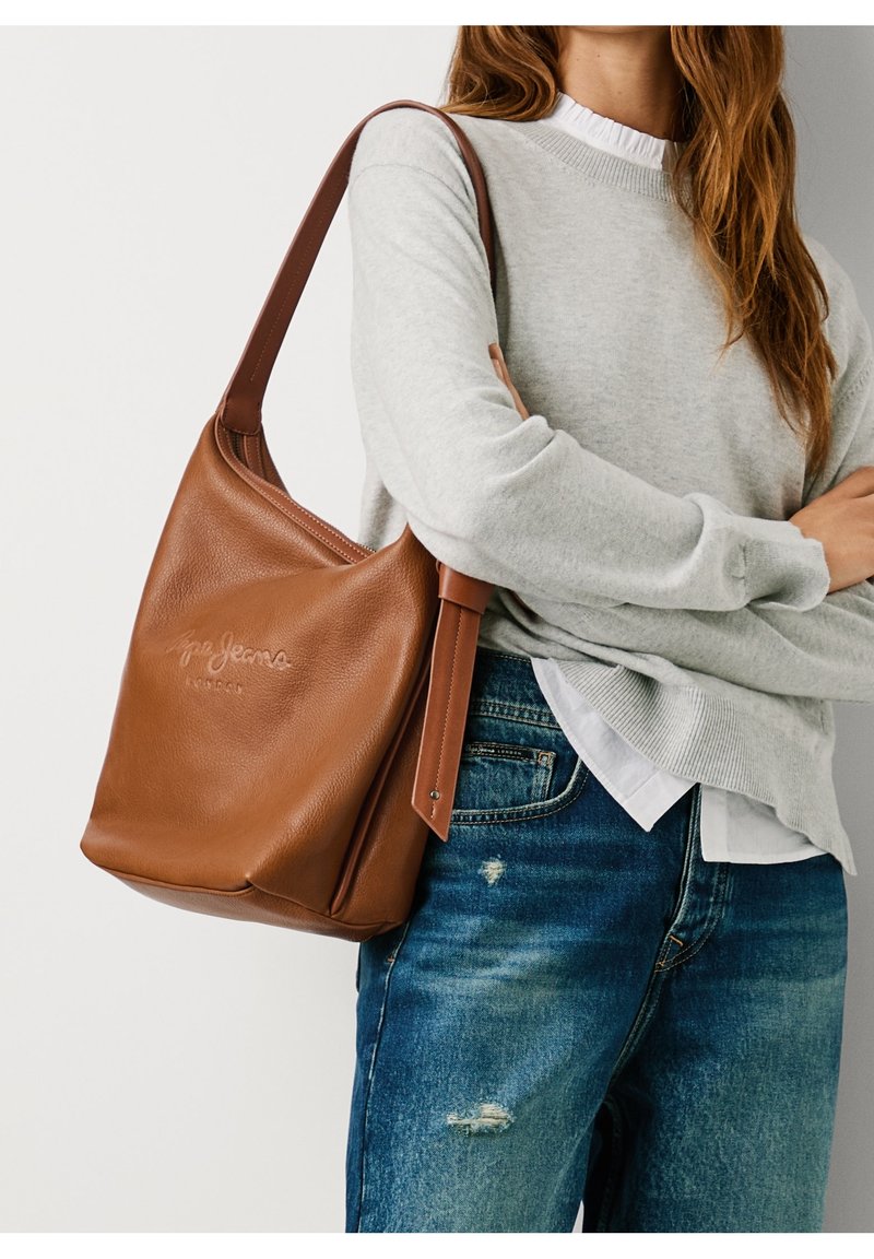 Sac à bandoulière en cuir marron porté sur un pull gris et un jean bleu délavé sur une personne aux bras croisés.