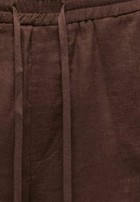 Pantalon en tissu marron avec une taille élastique et de longs cordons pendants, montrant une poche latérale sur le côté gauche.