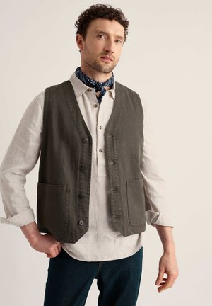 Uomo che indossa un gilet scuro abbottonato sopra una camicia chiara a maniche lunghe con polsini arrotolati e una sciarpa decorata al collo.
