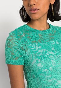 Top de renda verde de mangas curtas com padrões florais intrincados e decote redondo. O tecido é transparente com um acabamento texturizado.