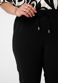 Pantalons à taille élastique noire avec une poche latérale, en tissu texturé, et fermeture à cordon de serrage avec embouts métalliques argentés.