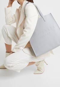 Šedá kožená taška přes rameno s embosovaným logem "Ted Baker London". Má ploché uchy a hladký povrch. Modelka má na sobě světle zbarvený outfit.