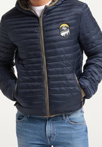 Doudoune marine avec un design matelassé horizontal, fermeture éclair avant et poches latérales. Caractérisée par un badge logo sur le côté gauche de la poitrine et une capuche.