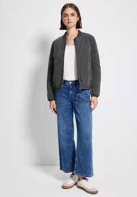 Giacca bomber grigia con zip, polsini e vita elastici, abbinata a jeans a gamba larga di un blu chiaro e sneakers bianche. Design semplice e moderno.