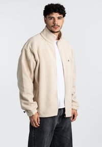 Giacca di pile beige con colletto alto, parte anteriore con zip, spalle scese e polsini elasticizzati. Indossata sopra una camicia bianca e jeans grigi.