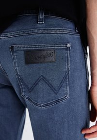 Donkerblauwe denimjeans met een zwart leren "Wrangler" label op de achterzak, met kenmerkende stiksels in een zigzagpatroon.