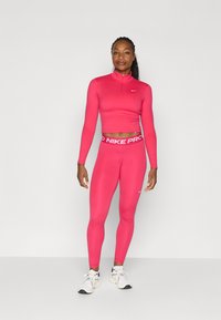 Tætsiddende lyserød atletisk crop top med høj krave, parret med matchende lyserøde leggings med en hvid "NIKE PRO" talje. Hvide sneakers.