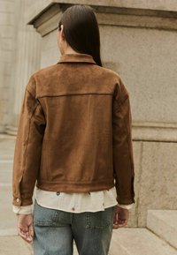 Braune Wildlederjacke mit Kragen, sorgfältig verarbeiteten Nähten und Knopfmanschetten, getragen über einem leichten, bestickten Hemd. Die Rückansicht zeigt die Passform der Jacke.