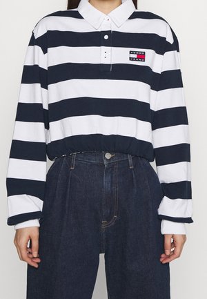 Persoon draagt een lange mouwen, zwart-wit horizontaal gestreept poloshirt met "Tommy Jeans"-logo en hoog getailleerde donkere spijkerbroek.