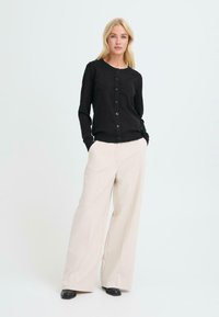 Cardigan nero con scollo rotondo e apertura frontale con bottoni, abbinato a pantaloni larghi crema e scarpe nere su uno sfondo chiaro.