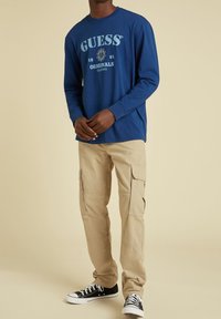 Blaues Langarmhemd mit "GUESS Originals 1981" Grafik, kombiniert mit beigen Cargo-Hosen mit Taschen und schwarzen Sneakers mit weißen Sohlen.