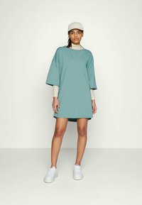 Mulher vestindo um vestido oversized em teal sobre uma blusa de manga longa, chapéu branco, tênis e colar, de pé contra um fundo claro e simples.