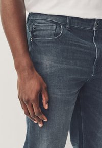 Jeans délavés foncés portés par une personne à la peau foncée, montrant la poche avant et une main reposant sur la cuisse sur un fond clair.