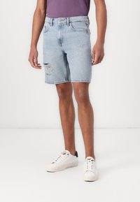 Shorts in denim azzurro con design strappato, orlo sfilacciato e stile a cinque tasche, abbinati a sneakers bianche.