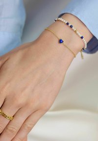 Bracciale in oro con un ciondolo a forma di cuore blu, accompagnato da un bracciale con perle e perle blu, indossato su una mano con un anello d'oro.