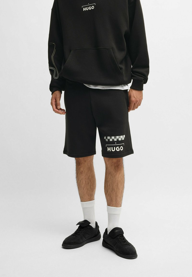 Haut à capuche noir avec logo blanc et contour contrasté, associé à un short noir avec des accents à carreaux et un logo. Baskets noires et chaussettes blanches.