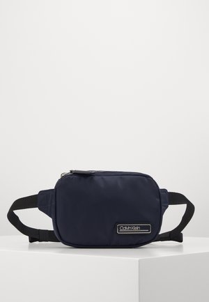 Borsa a vita rettangolare blu navy con cinturini neri regolabili e una placca con il logo Calvin Klein sul davanti, posizionata su una superficie bianca.