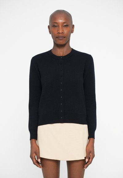 Un cardigan en bleu marine avec un col rond, des manches longues, une fermeture à boutons sur le devant et une texture lisse, associé à une jupe beige.