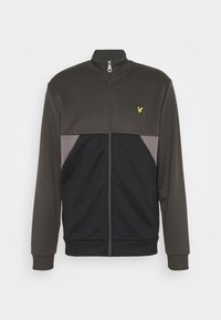 Chaqueta de cierre en color verde oscuro y negro con cuello alto, que presenta paneles laterales grises y un logotipo amarillo en la parte superior izquierda del pecho.