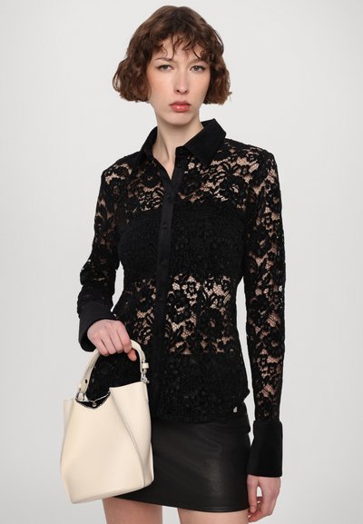 Svart bluse i blonder med en solid svart krage og mansjetter, med blomster mønster. Modellen holder en kremfarget bøtteveske med strukturert design.