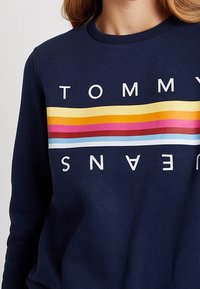 Marinblå sweatshirt med horisontella ränder i gult, orange, rosa, rött och blått, med vit text i versaler "TOMMY JEANS" ovanför och nedanför ränderna.