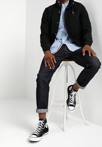 Veste bombardier noire sur une chemise bleue clair à boutons, jean denim foncé et baskets montantes noires avec accents blancs, assis sur un tabouret.