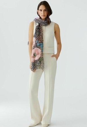 Vrouw in mouwloze crèmekleurige vest en broek met hand in de zak, draagster van een sjaal met luipaardprint en grote roze en blauwe bloemen.