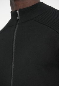 Čierny sveter na zips s vysokým golierom a rebrovanou textúrou na ramenách, s hladkým povrchom na tele.