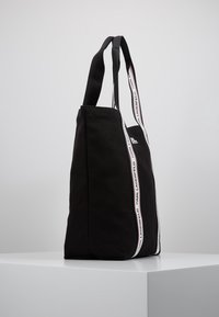 Černá plátěná taška na rameno s dvojitými černými uchy, vybavená svislým zipem a kontrastními bílými pásky s logem podél stran.