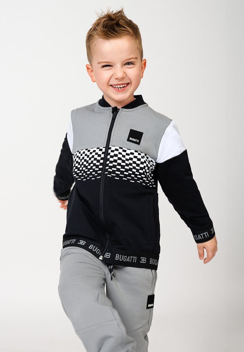 Schwarze und graue Zip-Jacke mit Schachbrettmuster. Ein Paar hellgraue Sweatpants mit einem schwarzen Logo-Bund.