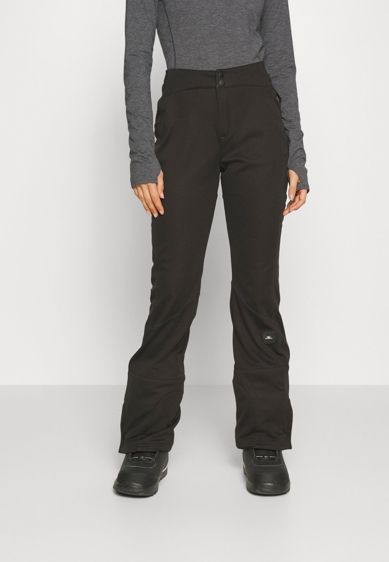 Kobieta ubrana w czarne spodnie narciarskie, czarne zimowe buty i dopasowaną szaro-niebieską koszulę z długim rękawem, stojąca na jednolitym białym tle.