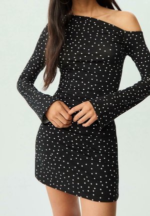 Woman wearing a black, long-sleeve, off-shoulder mini dress with white polka dots, hands clasped at waist.