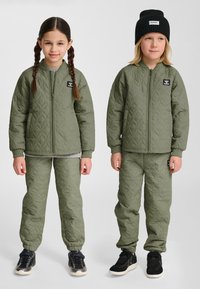 Dwoje dzieci ubranych w pasujące pikowane oliwkowo-zielone kurtki i spodnie, jedno z warkoczami, drugie w czarnej czapce.