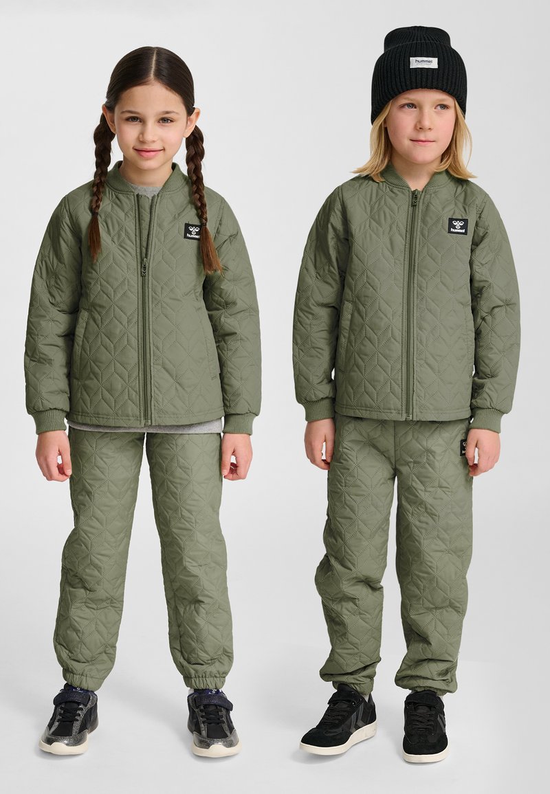 Dwoje dzieci ubranych w pasujące pikowane oliwkowo-zielone kurtki i spodnie, jedno z warkoczami, drugie w czarnej czapce.