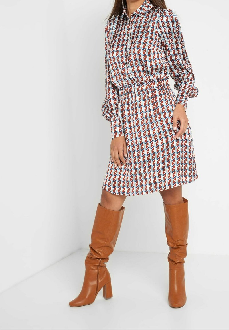 Femme portant une robe à motif géométrique à manches longues et des bottes marron en cuir à talons hautes jusqu'au genou sur un fond blanc.