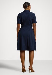 Žena v námornícky modrých krátkych rukávoch a opaskom, v hnedých vysokých podpätkoch, stojaca chrbtom k pozorovateľovi na jednoduchom pozadí.