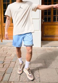Camiseta beige con un logo negro, pantalones cortos azules con puños estampados, calcetines blancos y zapatillas marrones con detalles crema. Entorno al aire libre.