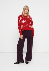 Pull en maille rouge avec des motifs de flocon de neige blancs, associé à un pantalon large violet et des chaussures à talons blocs noires.