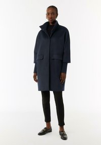 Marineblauer Wollmischmantel mit hohem Kragen, ausgestattet mit zwei Vorderaschen und geradem Schnitt. Kombiniert mit schwarzen, figurbetonten Hosen und Slippern.