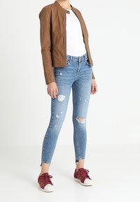 Giacca in finta pelle marrone, t-shirt bianca, jeans skinny blu strappati, sneakers rosse con suole bianche e dettagli dorati.