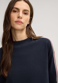Marineblauwe sweatshirt met een ronde hals, voorzien van contrasterende witte en bordeauxrode strepen langs de mouwen. Glad, textuurstof.