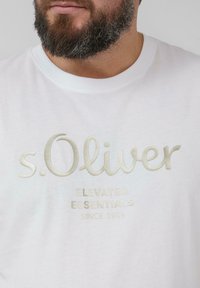 Weißes T-Shirt mit kurzen Ärmeln und metallic-goldener, gestickter Markenlogo "s.Oliver" sowie dem Text "ELEVATED ESSENTIALS SINCE 1969" darunter.